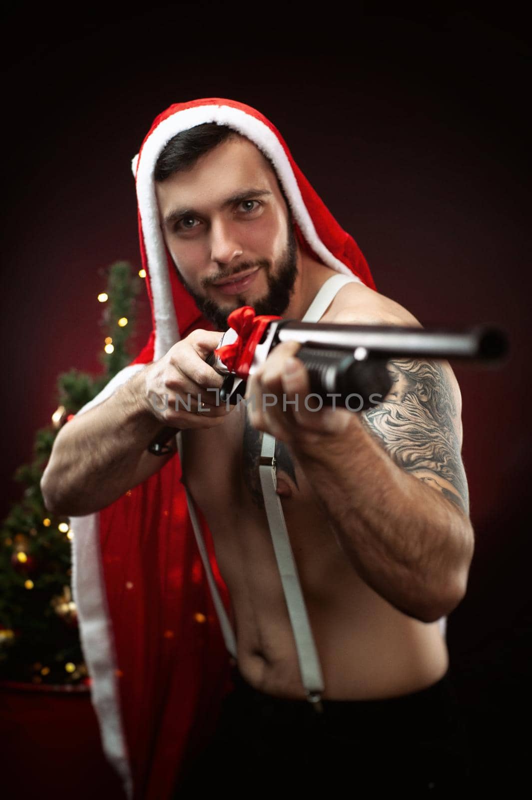 strong guy in a red Santa Claus cape at the Christmas tree with a shotgun in his hands