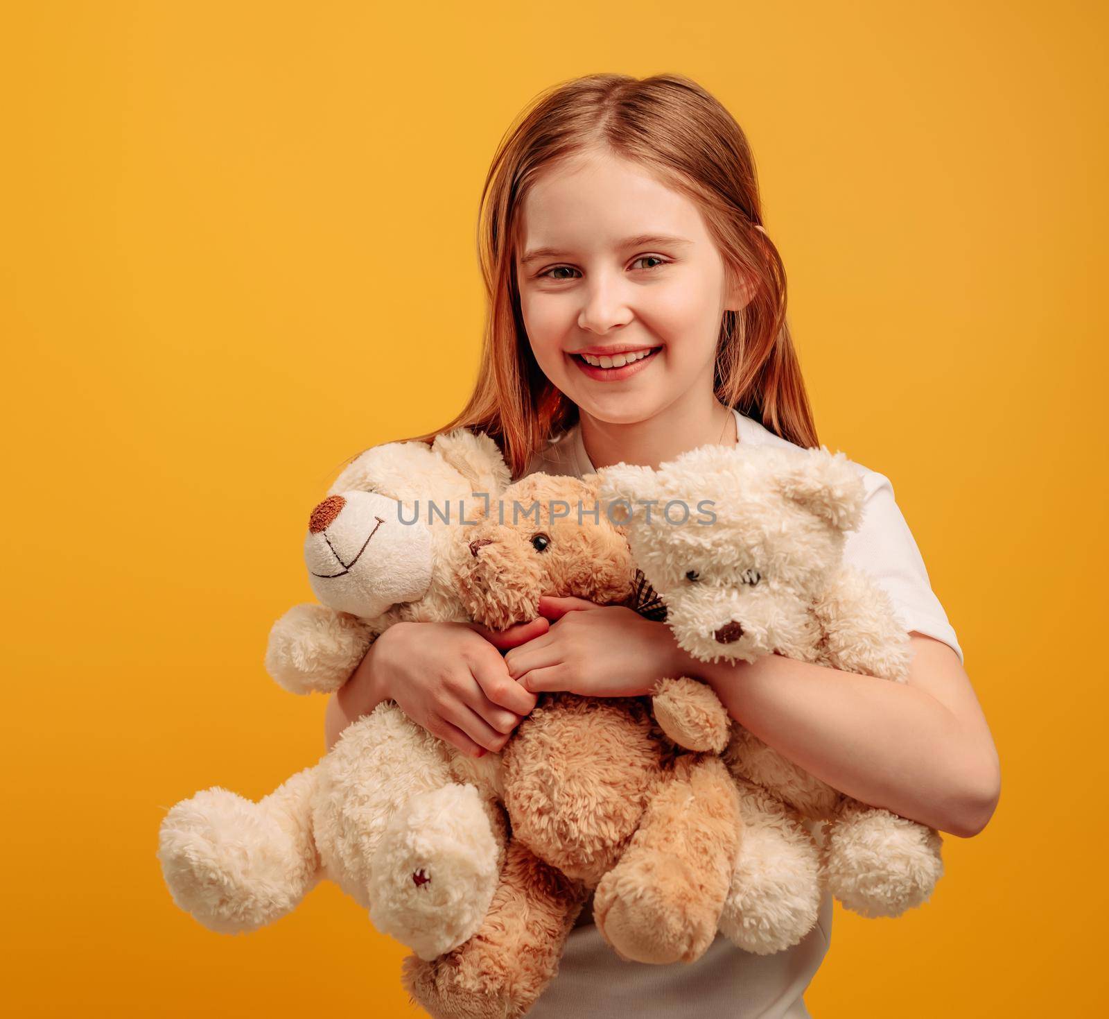 Beautiful girl child with three teddy bears isolated on yellow background with copyspace. Portrait of kid holding toys and smiling looking at the camera