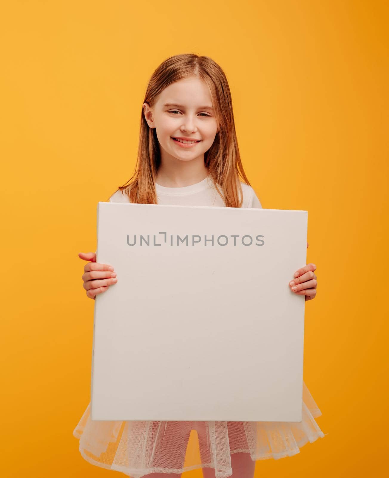 Beautiful girl child holding white canvas and looking at the camera isolated on yellow background with copyspace. Portrait of kid holding linen and smiling