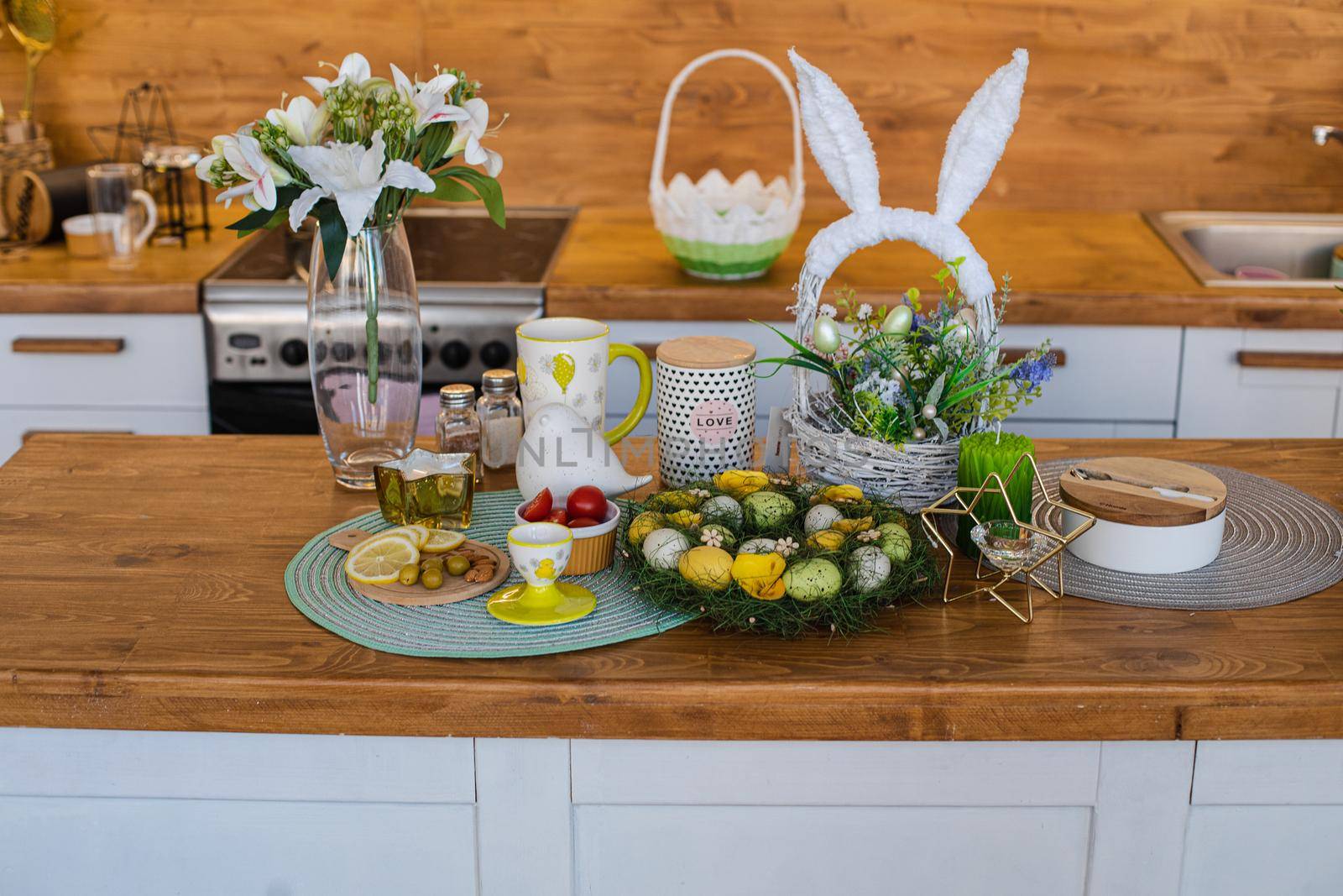 Still life of painted Easter eggs in wreath with lemon and olives and tomatoes on wooden kitchen counter. A lovely basket with bunny ears with flowers and decorative eggs. Vase of lilies on the counter.
