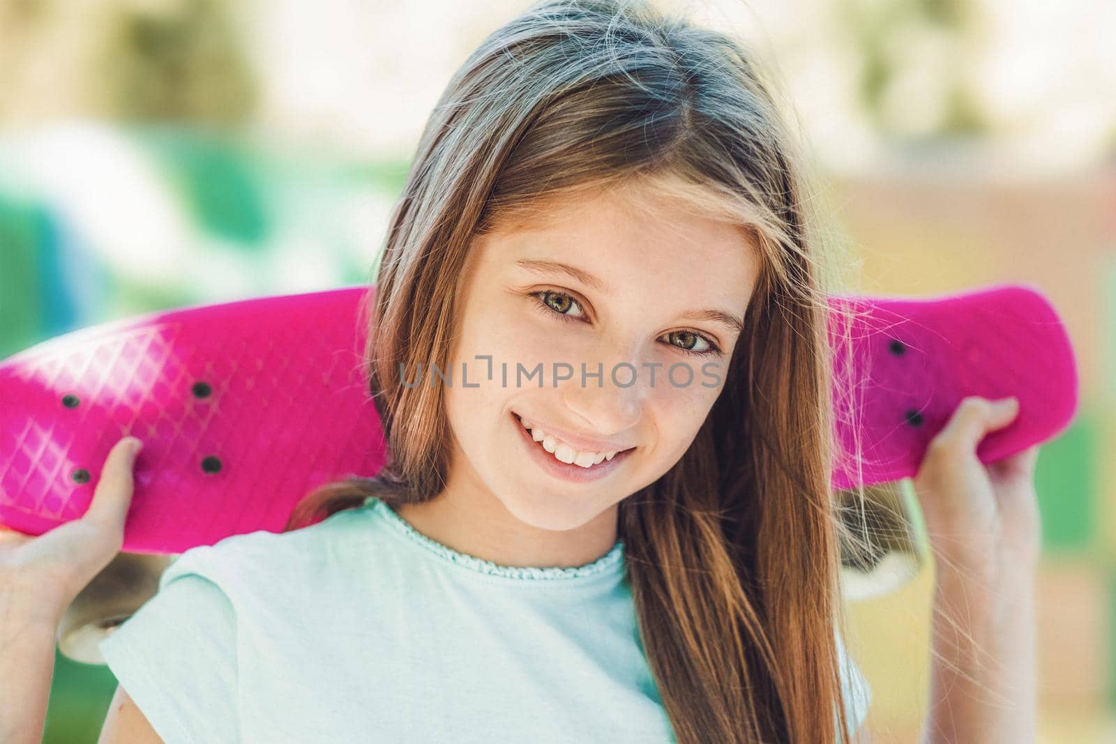 Closeup portrait of smiling teenage girl with pink skateboard behind her back in summertime