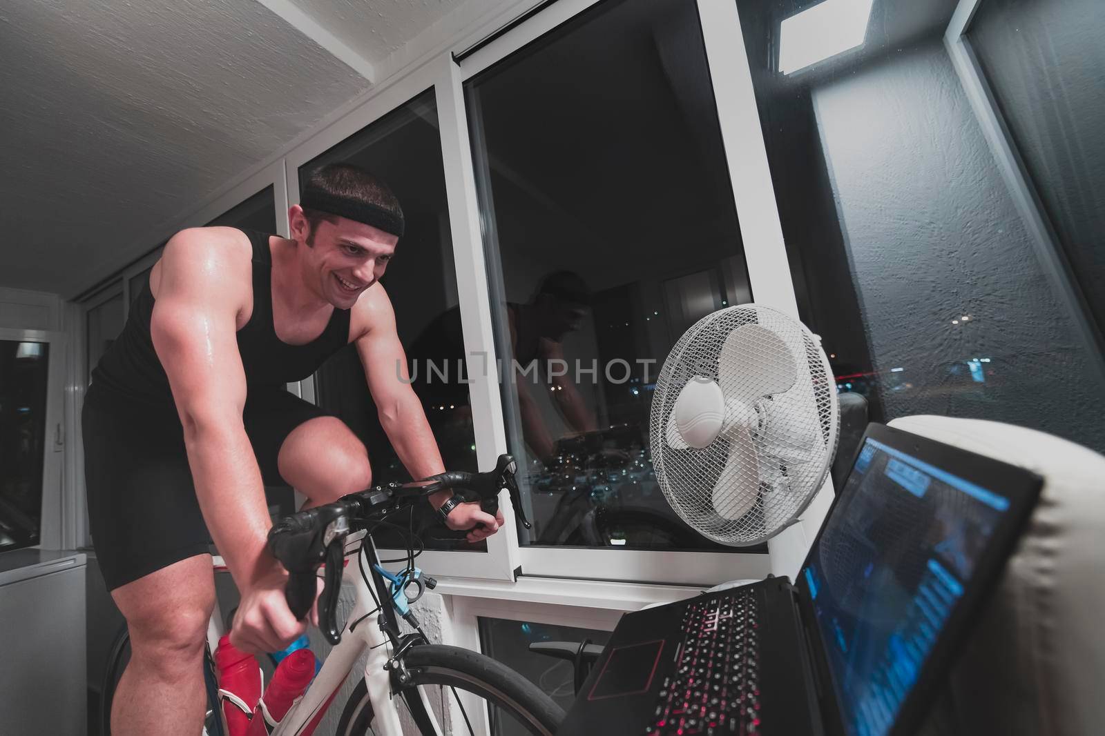 Man cycling on the machine trainer he is exercising in the home at night. Playing online bike racing game during coronavirus covid19 lockdown. New normal concept.