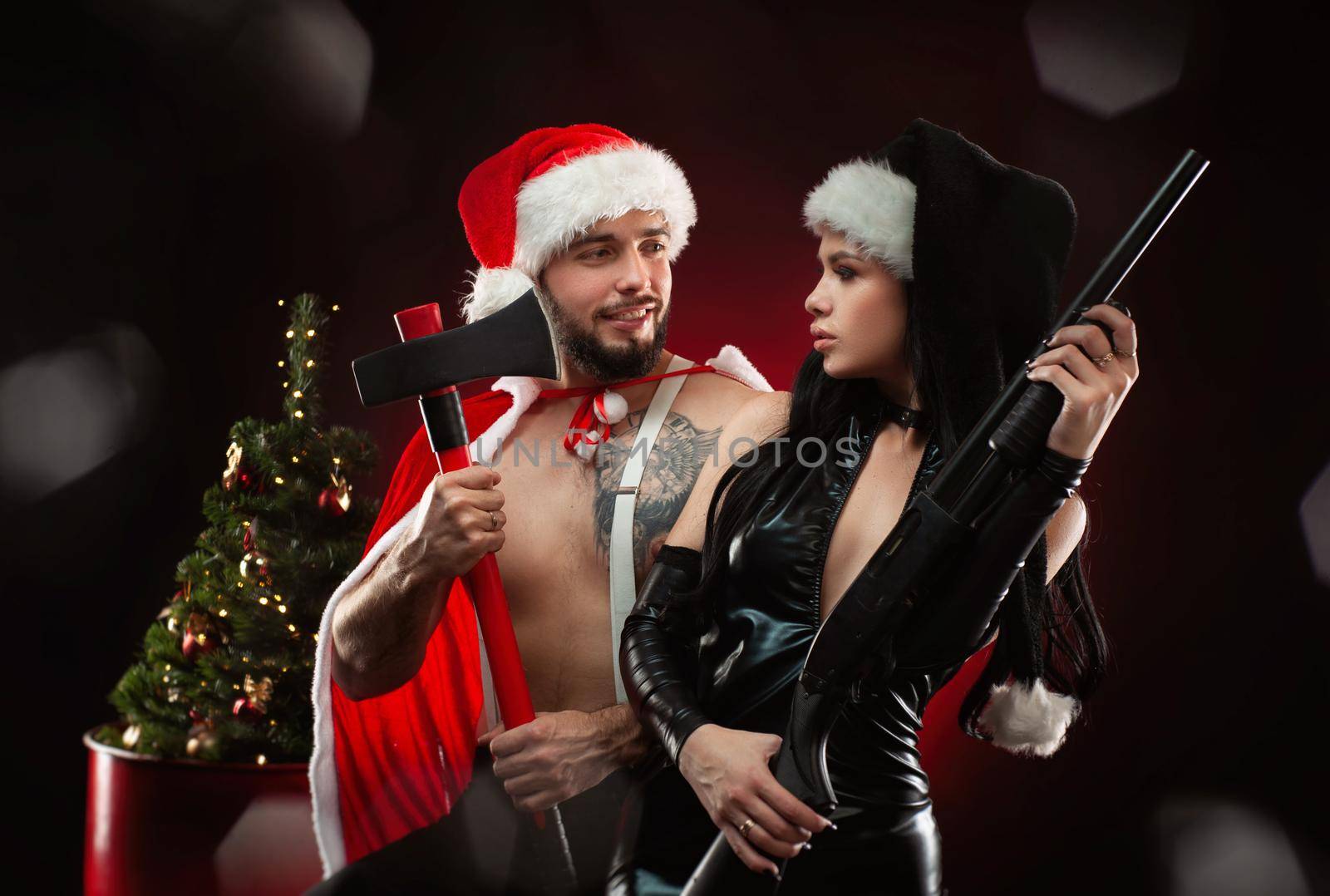 Christmas romantic photo shoot of a guy and a girl in New Year's Santa Claus costumes with weapons, a Christmas tree on a dark red background