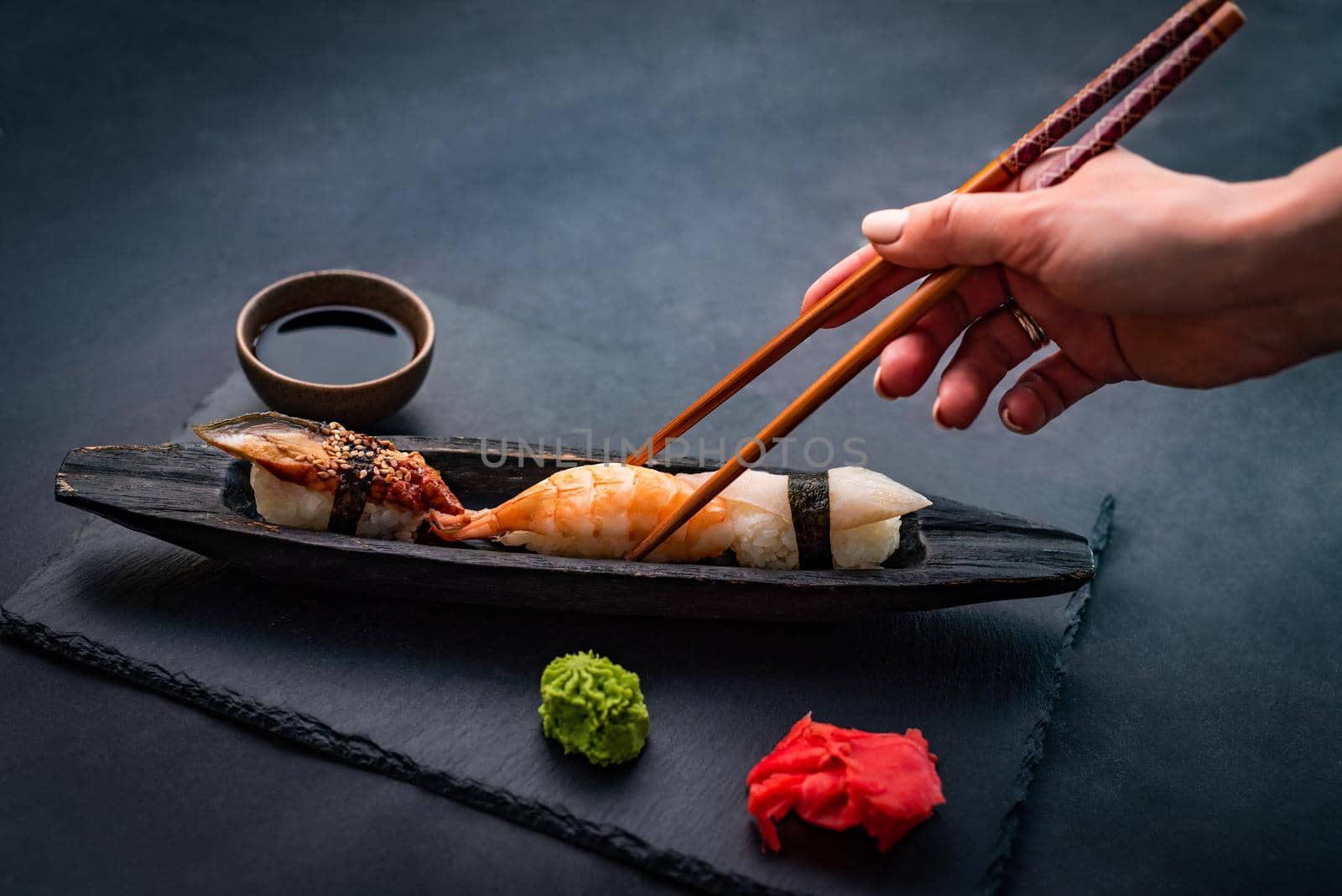Person hand holding chopsticks and eat sushi sashimi set with shrimps, ginger and soy sauce served with wasabi on black table. Traditional healthy japanese meal with rice and seafood