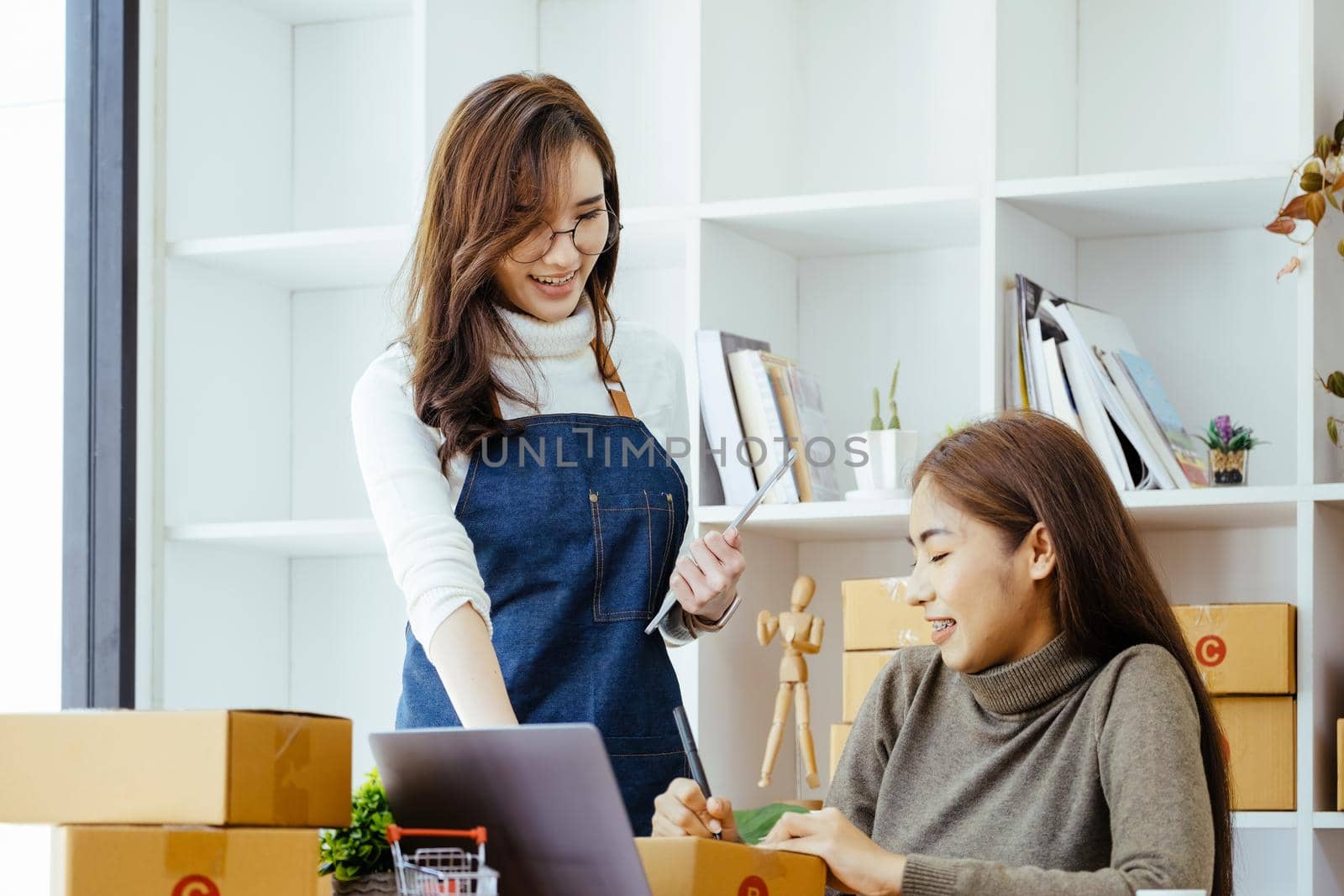 selling products online, the focus is on the face of a woman in white pointing to a friend to write down the shipping address for the customer and prepare it for postage. by Manastrong