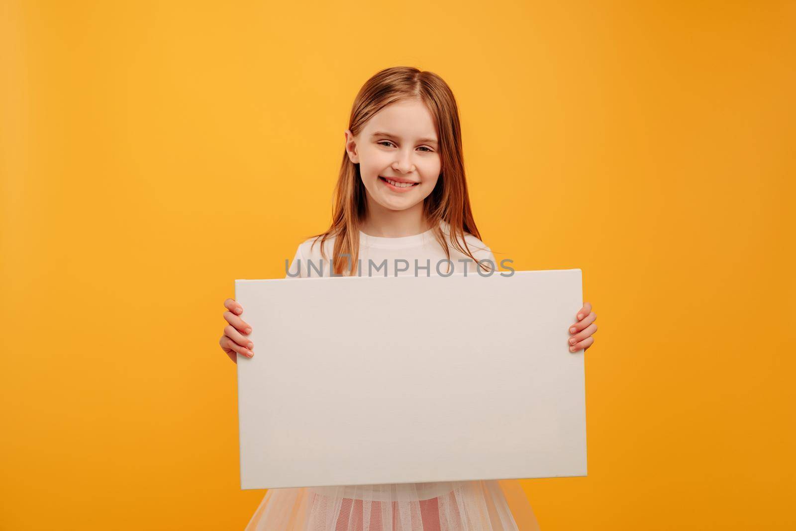 Pretty smiling girl child holding white canvas and looking at the camera isolated on yellow background with copyspace. Horizontal portrait of kid holding linen with place for text