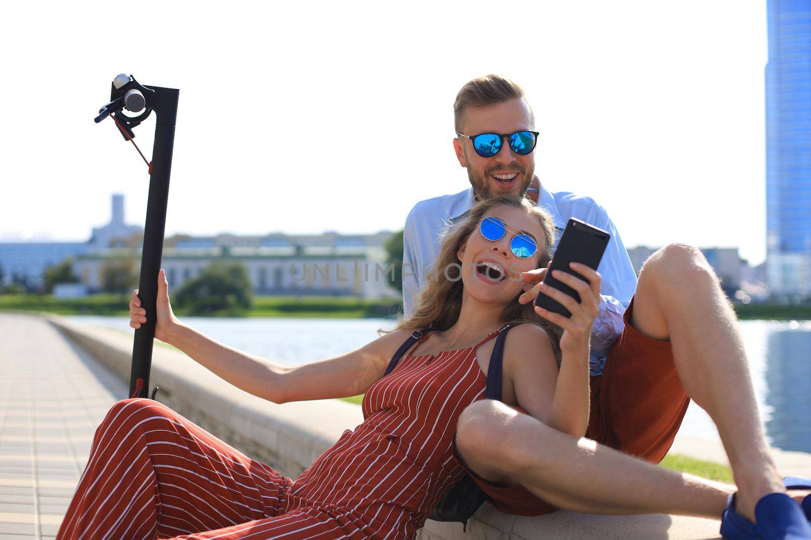 Lovely couple having fun driving electric scooter, taking a break from driving, sitting on riverbank, taking selfie by tsyhun