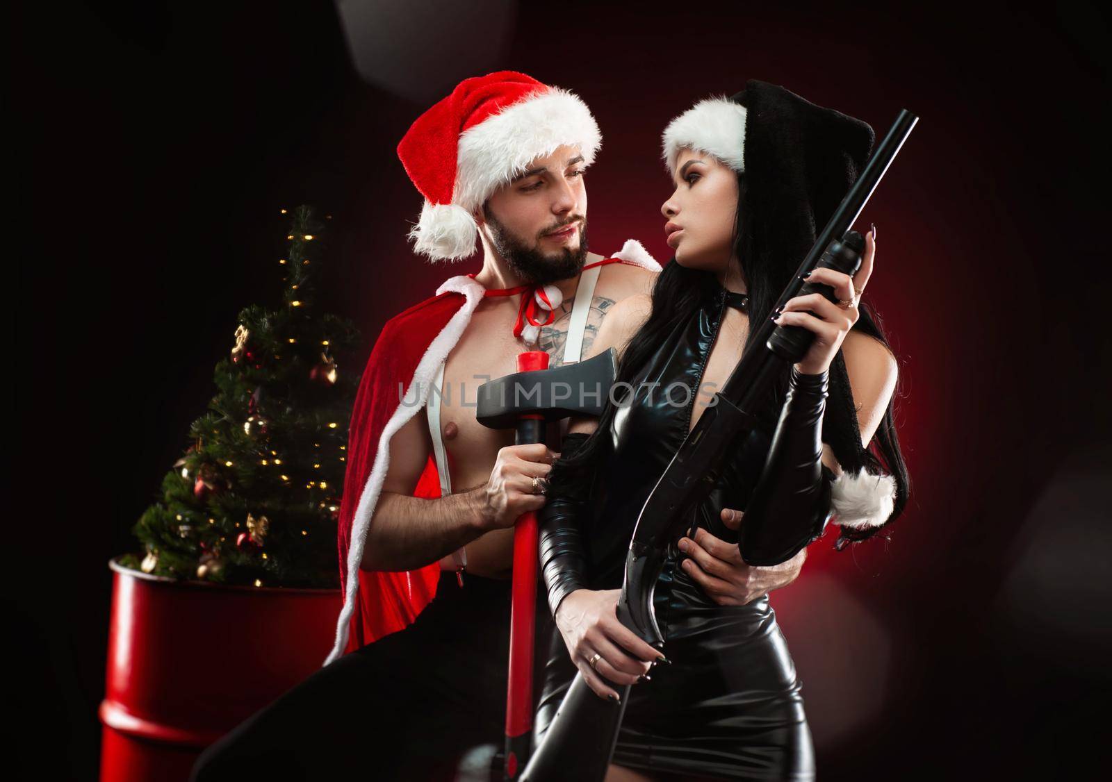 Christmas love photo shoot of a guy and a girl in New Year's Santa Claus costumes with weapons, a Christmas tree on a dark red background by Rotozey