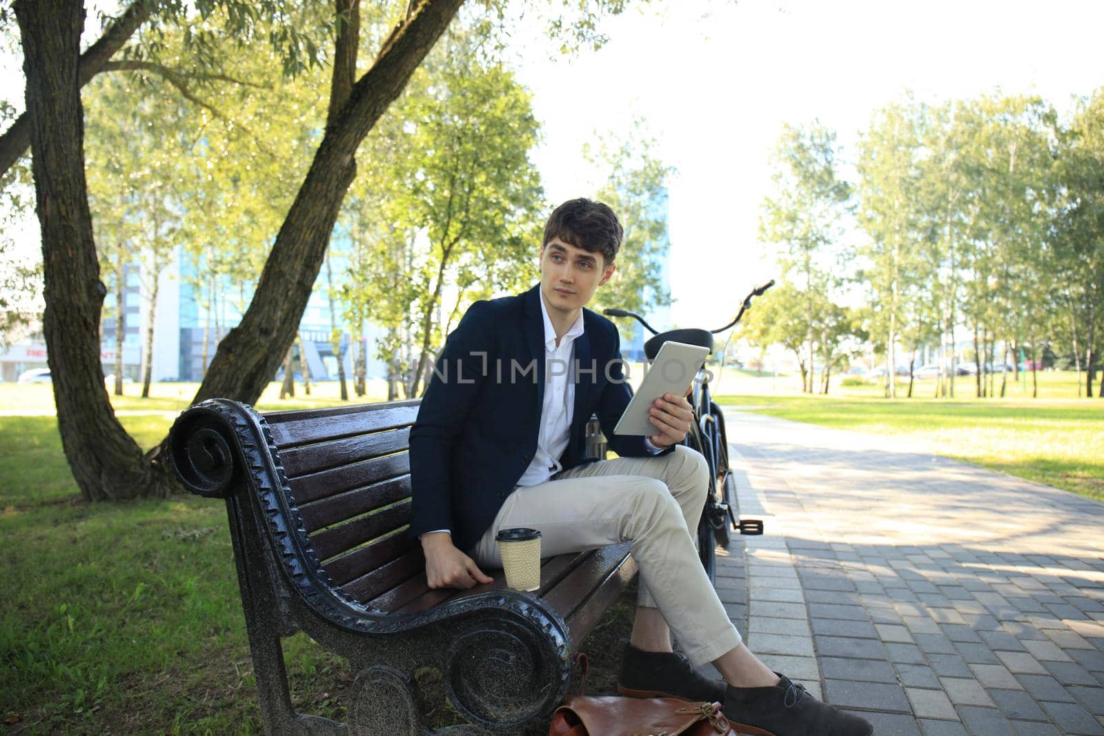 Businessman on a coffee break. He is sitting on a bench and working at touchpad, next to bike.