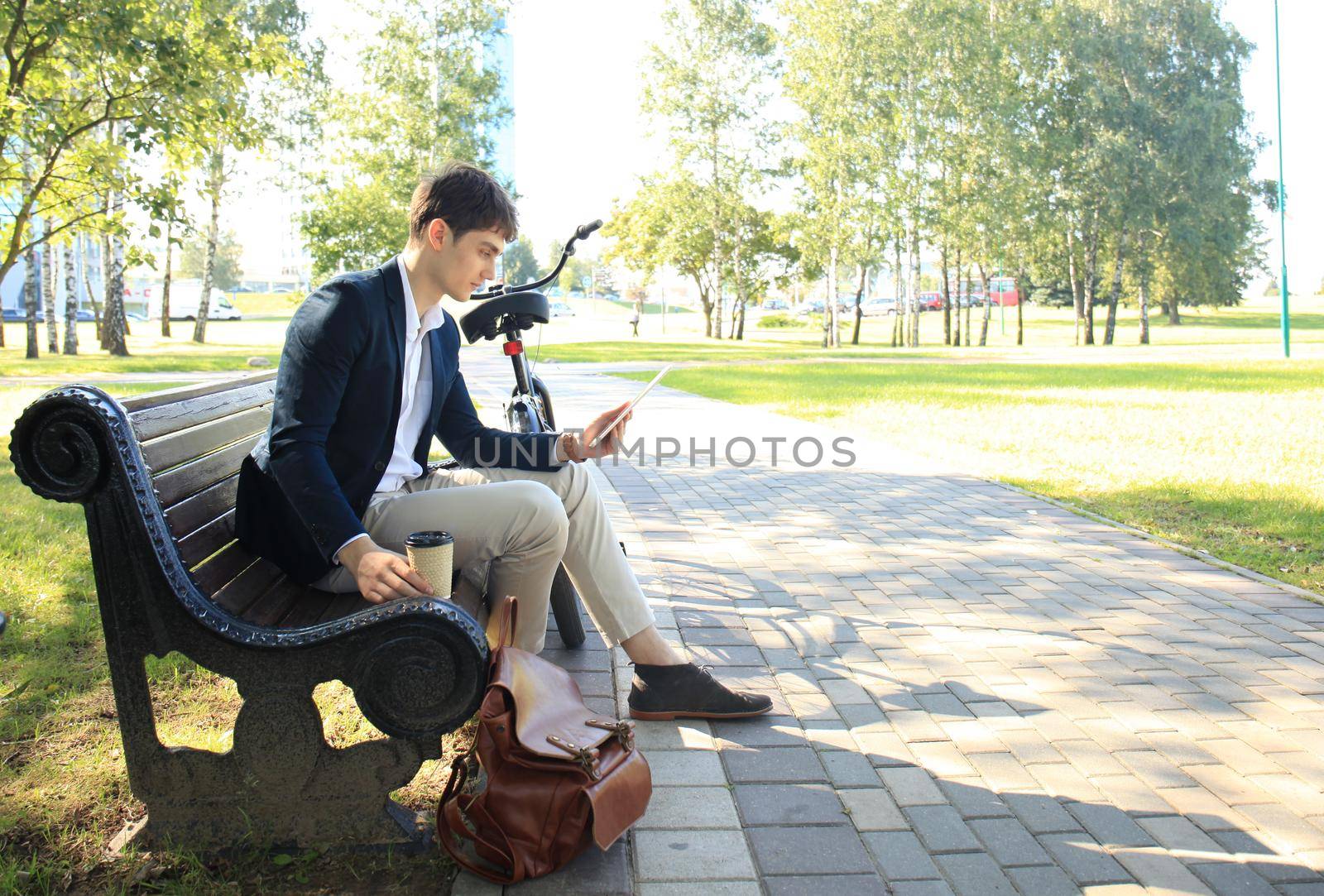 Businessman on a coffee break. He is sitting on a bench and working at touchpad, next to bike.