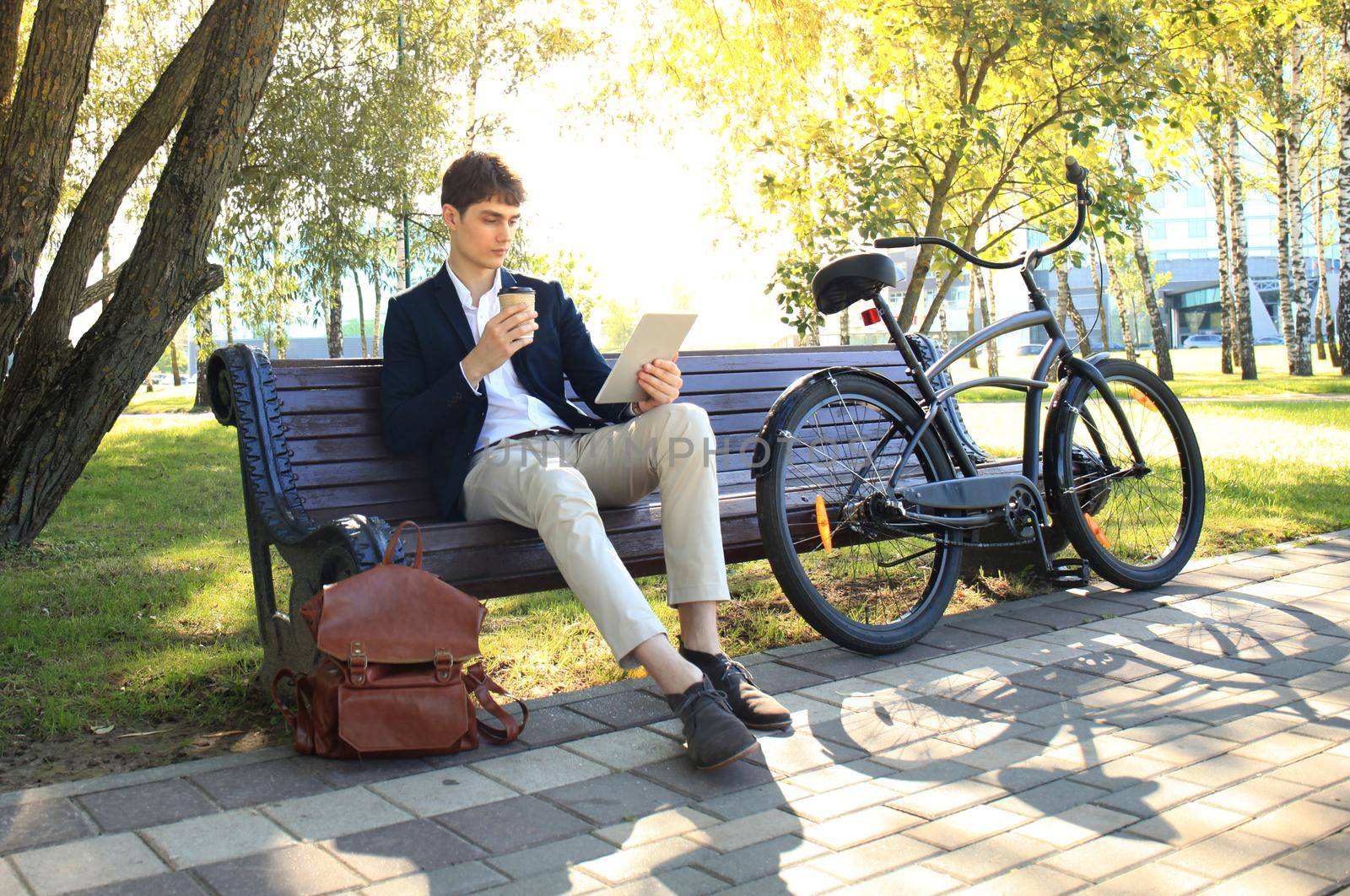 Businessman on a coffee break. He is sitting on a bench and working at touchpad, next to bike.