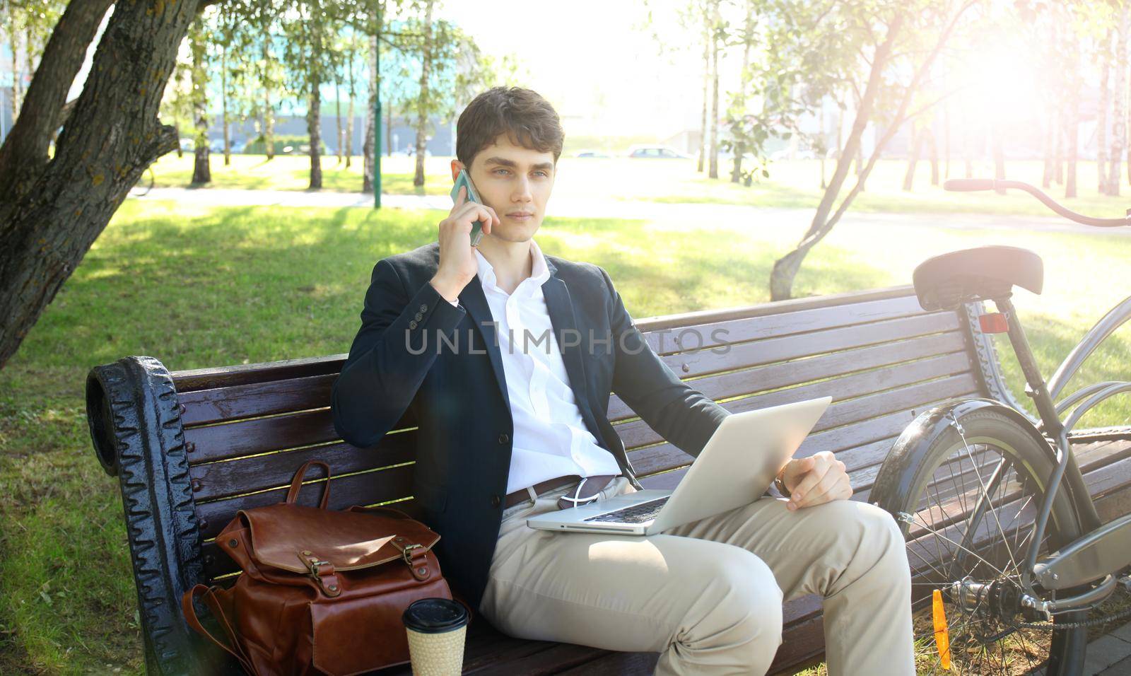 Businessman on a coffee break. He is sitting on a bench and working at laptop, next to bike.