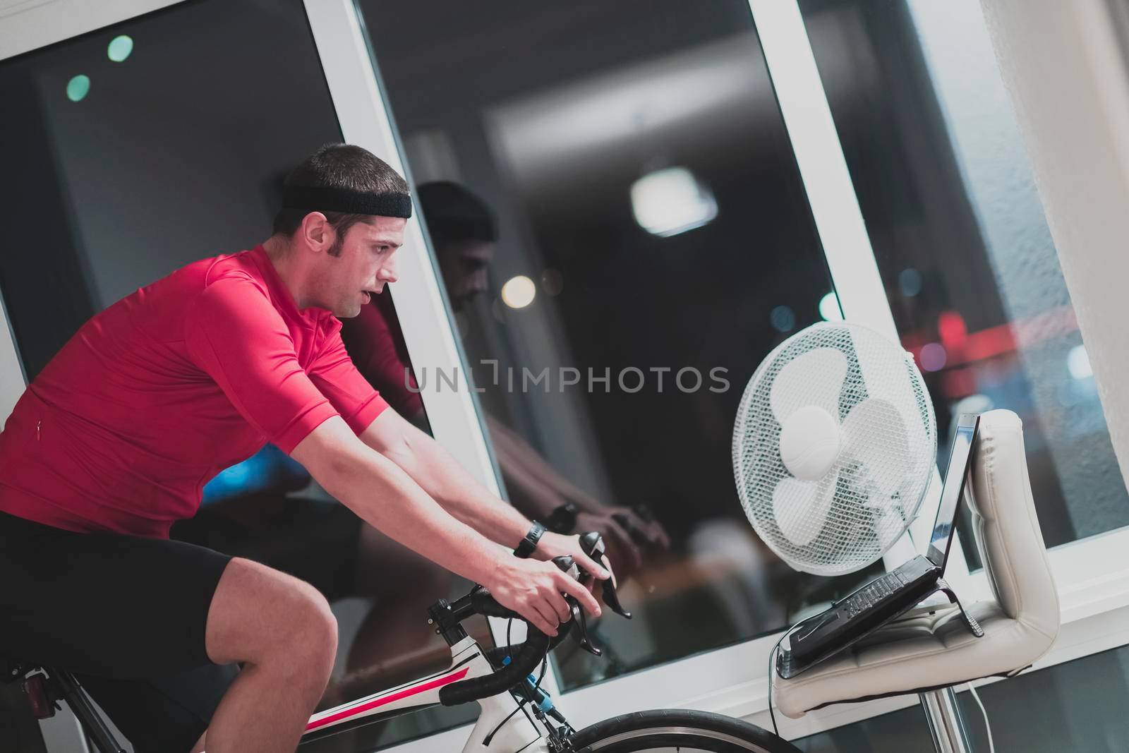 Man cycling on the machine trainer he is exercising in the home at night. Playing online bike racing game during coronavirus covid19 lockdown. New normal concept.