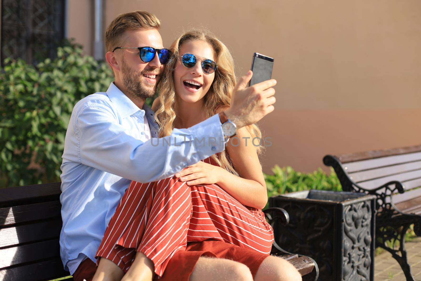 Romantic young couple in summer clothes smiling and taking selfie while sitting on bench in city street by tsyhun