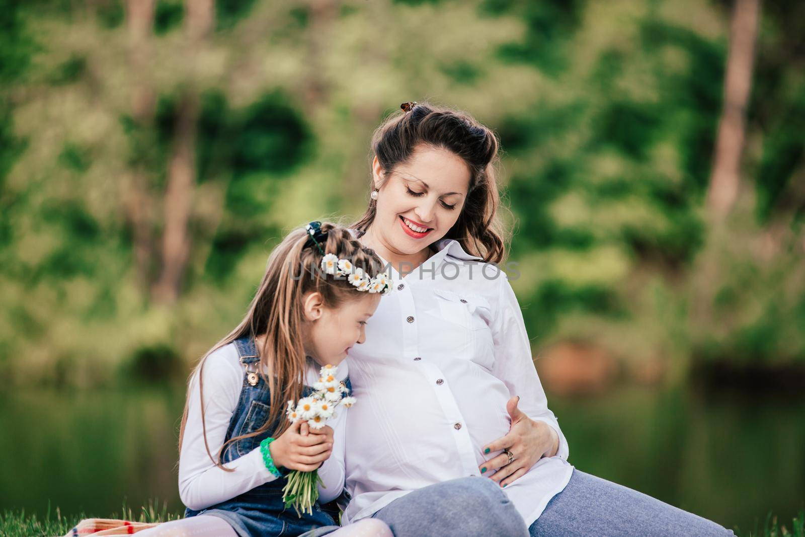 beautiful pregnant woman with her little daughter sitting on the lawn on a summer day.