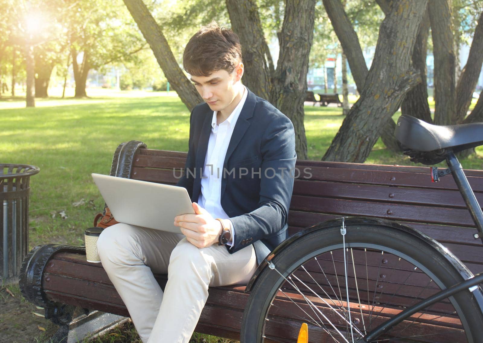 Businessman on a coffee break. He is sitting on a bench and working at touchpad, next to bike.