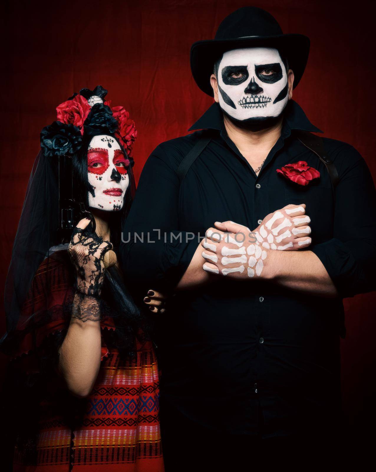 beautiful woman with a sugar skull makeup with a wreath of flowers on her head and a skeleton man in a black hat holding a gun by ndanko