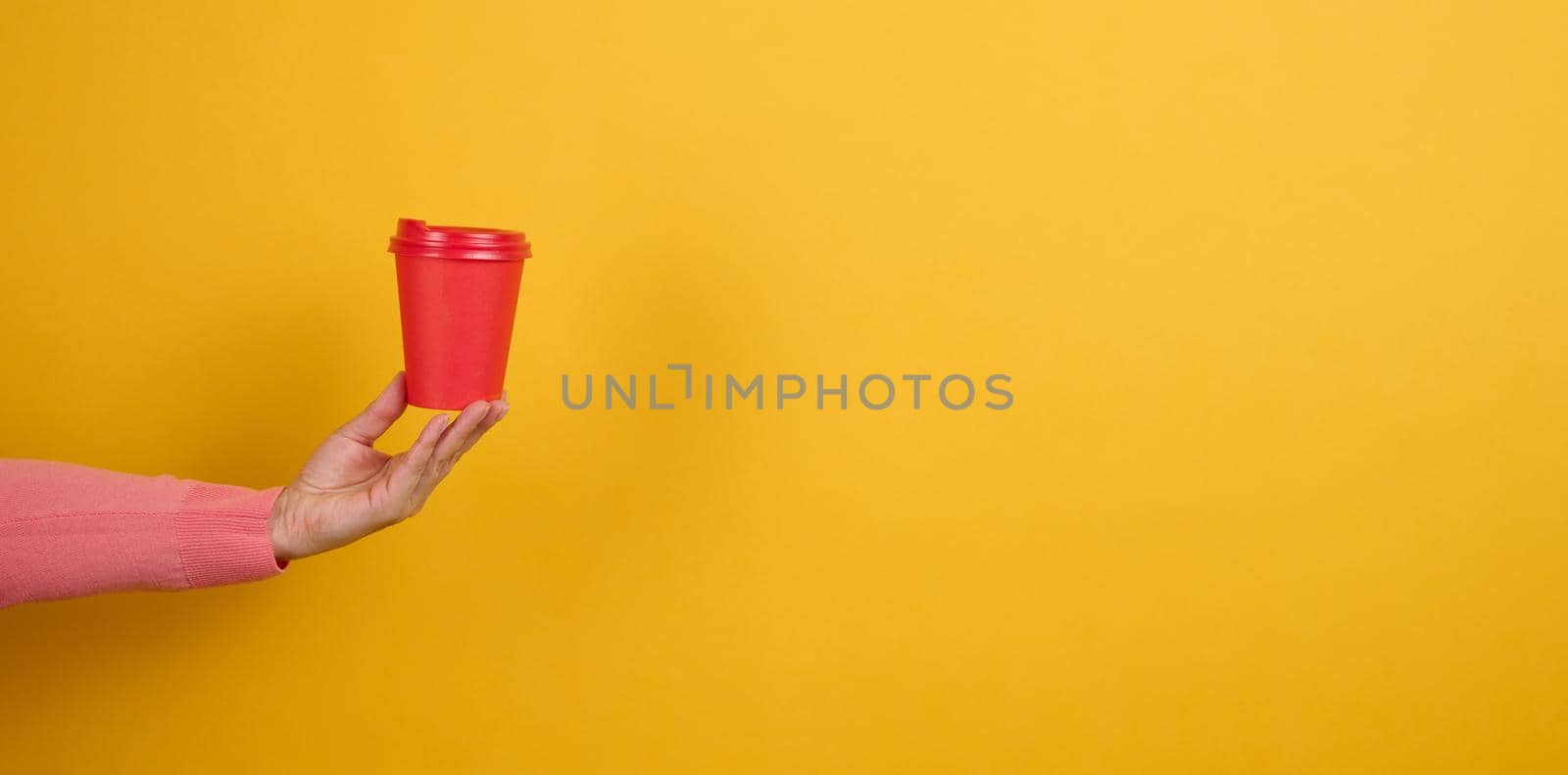 hand holds paper cardboard red cup for coffee, yellow background. Eco-friendly tableware, copy space by ndanko