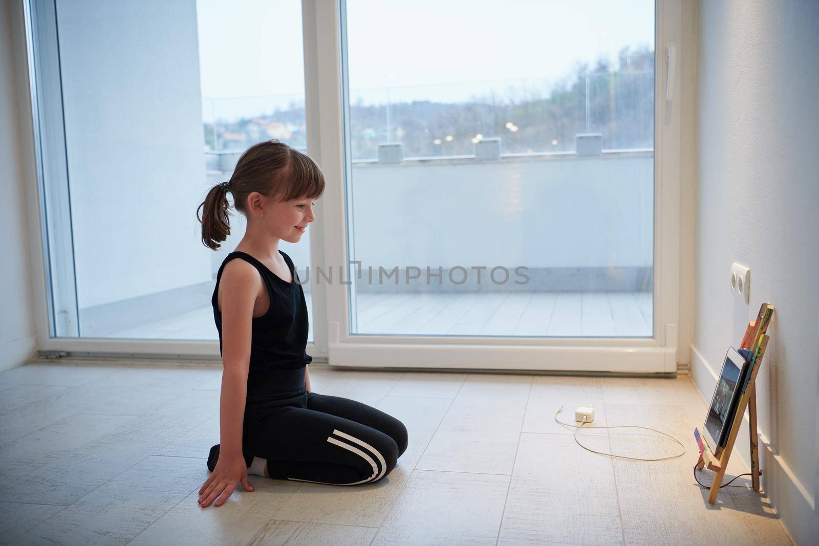 Little girl have online training while watching the tablet and doing fitness and modern ballet dance exercise at home. Online education concept class in modern ballet school. Social distance during quarantine, self-isolation in coronavirus pandemic staying at home time.