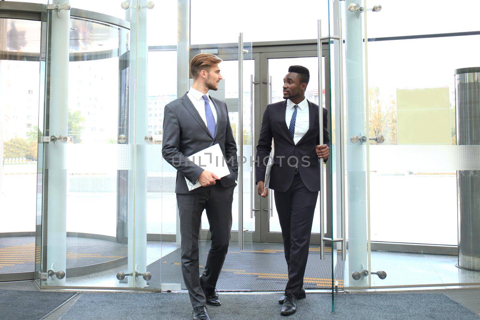 Two multinational young businessmen entering in office building with glass doors. by tsyhun