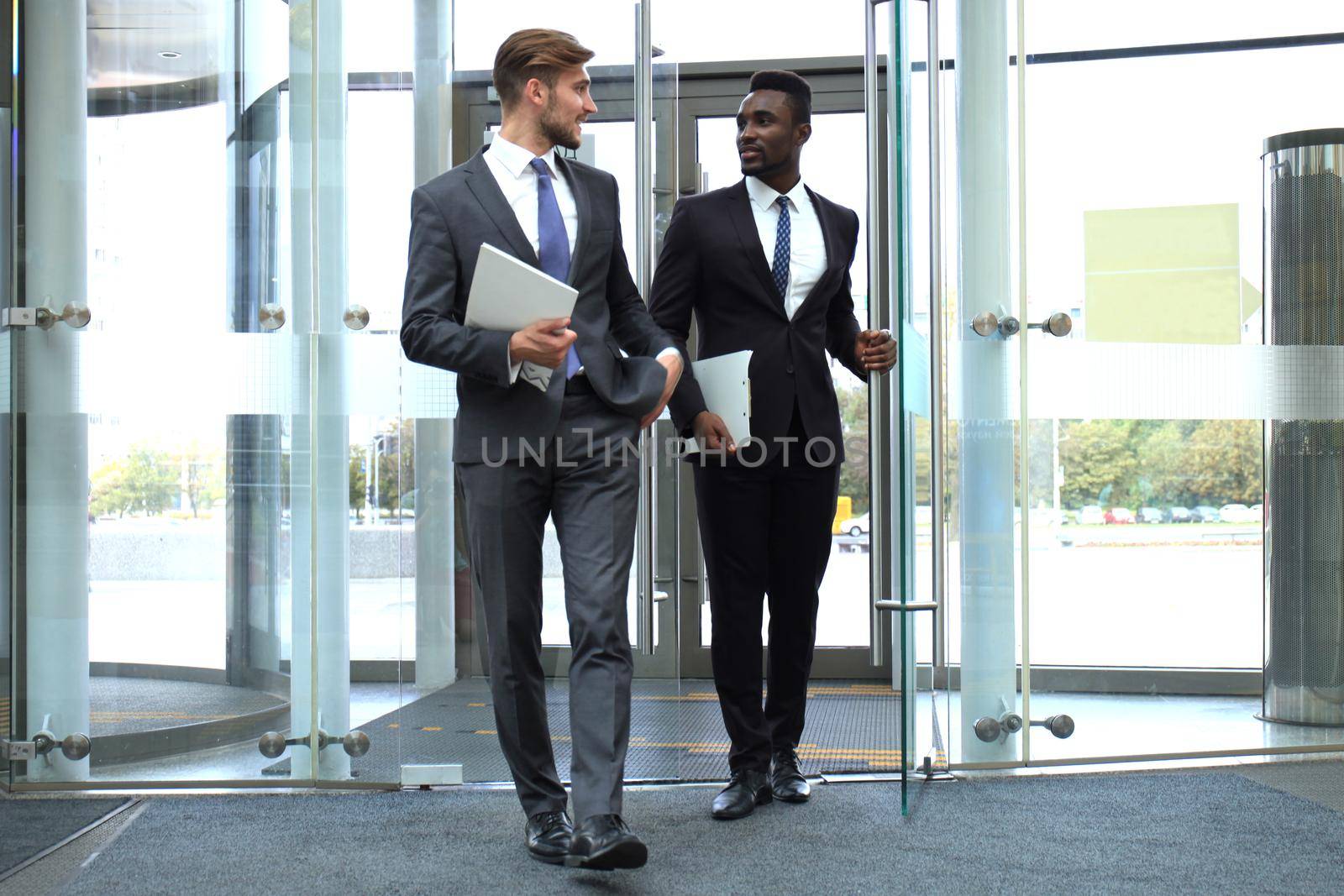 Two multinational young businessmen entering in office building with glass doors.