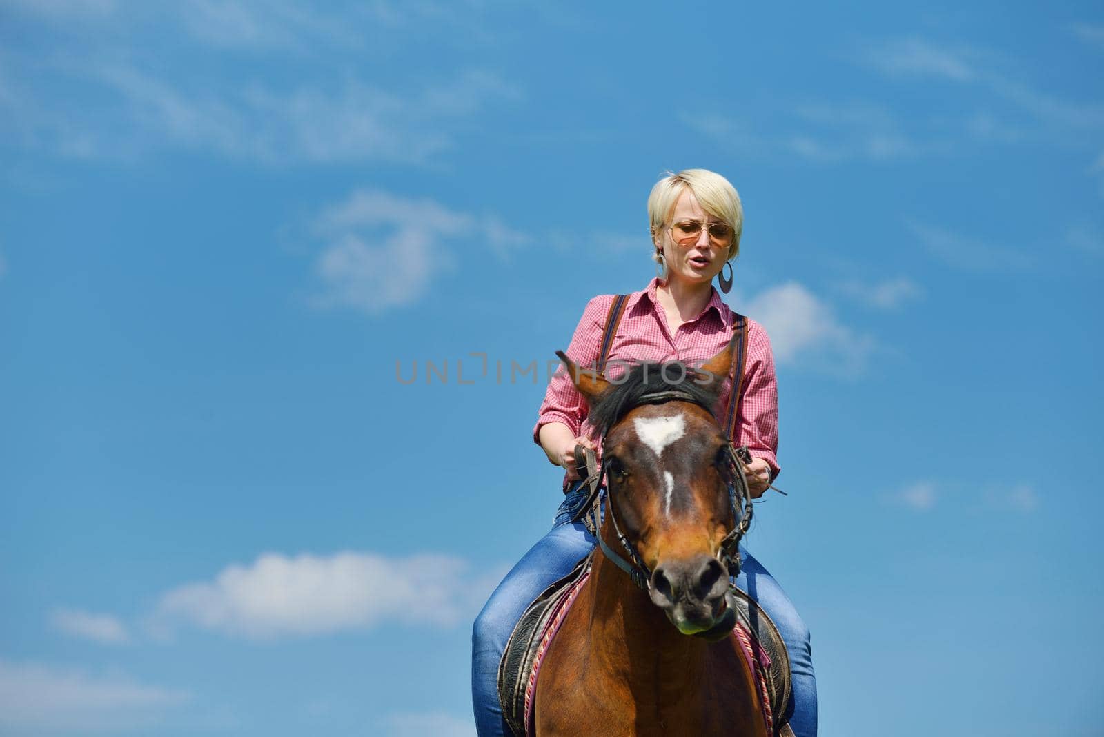 happy woman in sunglasses sitting on horse farm animal outdoors with blue sky in background