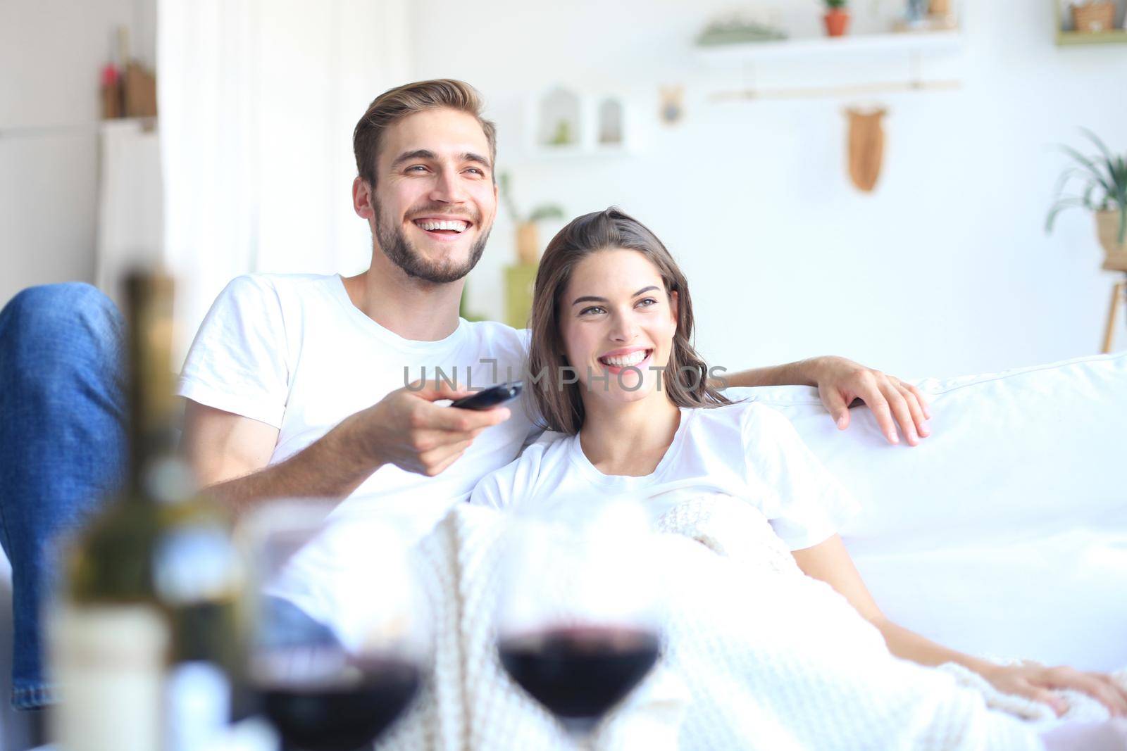 Young loving couple on sofa at home watching tv and laughing, drinking a glass of red wine. by tsyhun