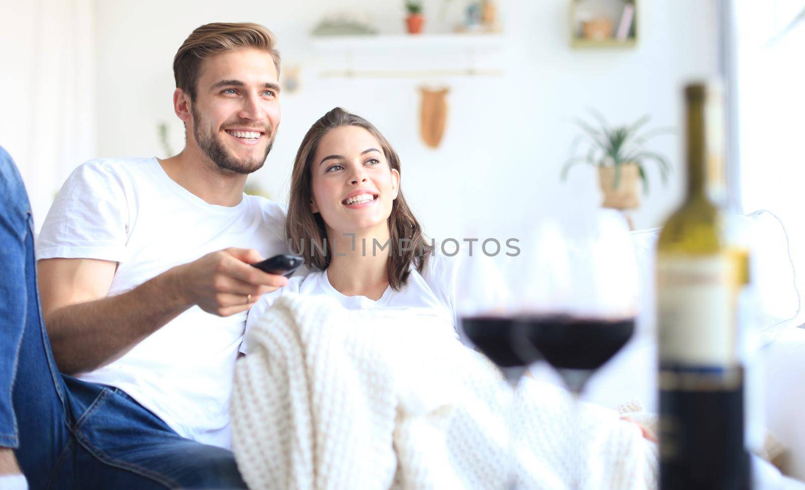 Young loving couple on sofa at home watching tv and laughing, drinking a glass of red wine. by tsyhun