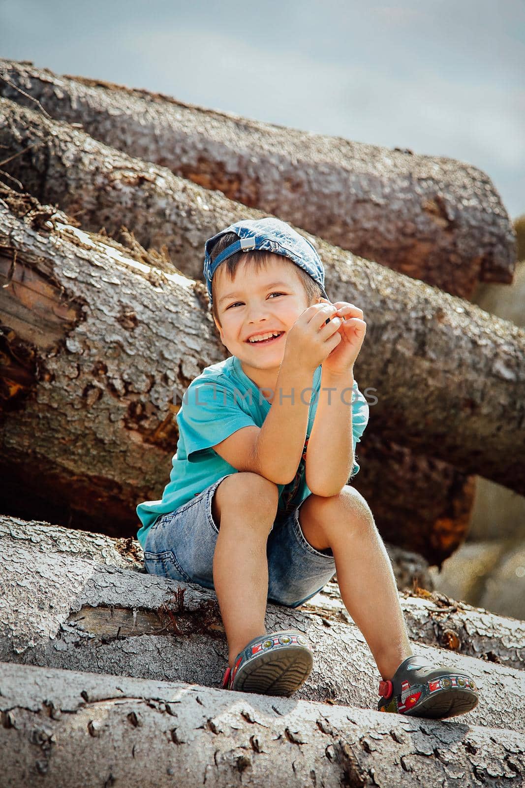 A small child in a blue baseball cap and short denim shorts sits on a log and laughs merrily