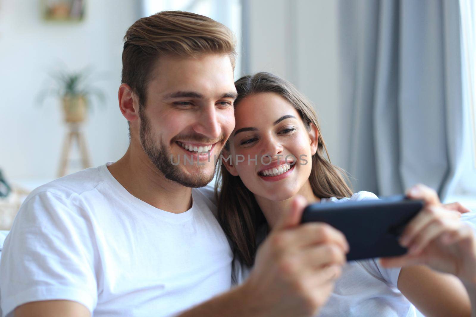 Young couple watching online content in a smart phone sitting on a sofa at home in the living room.