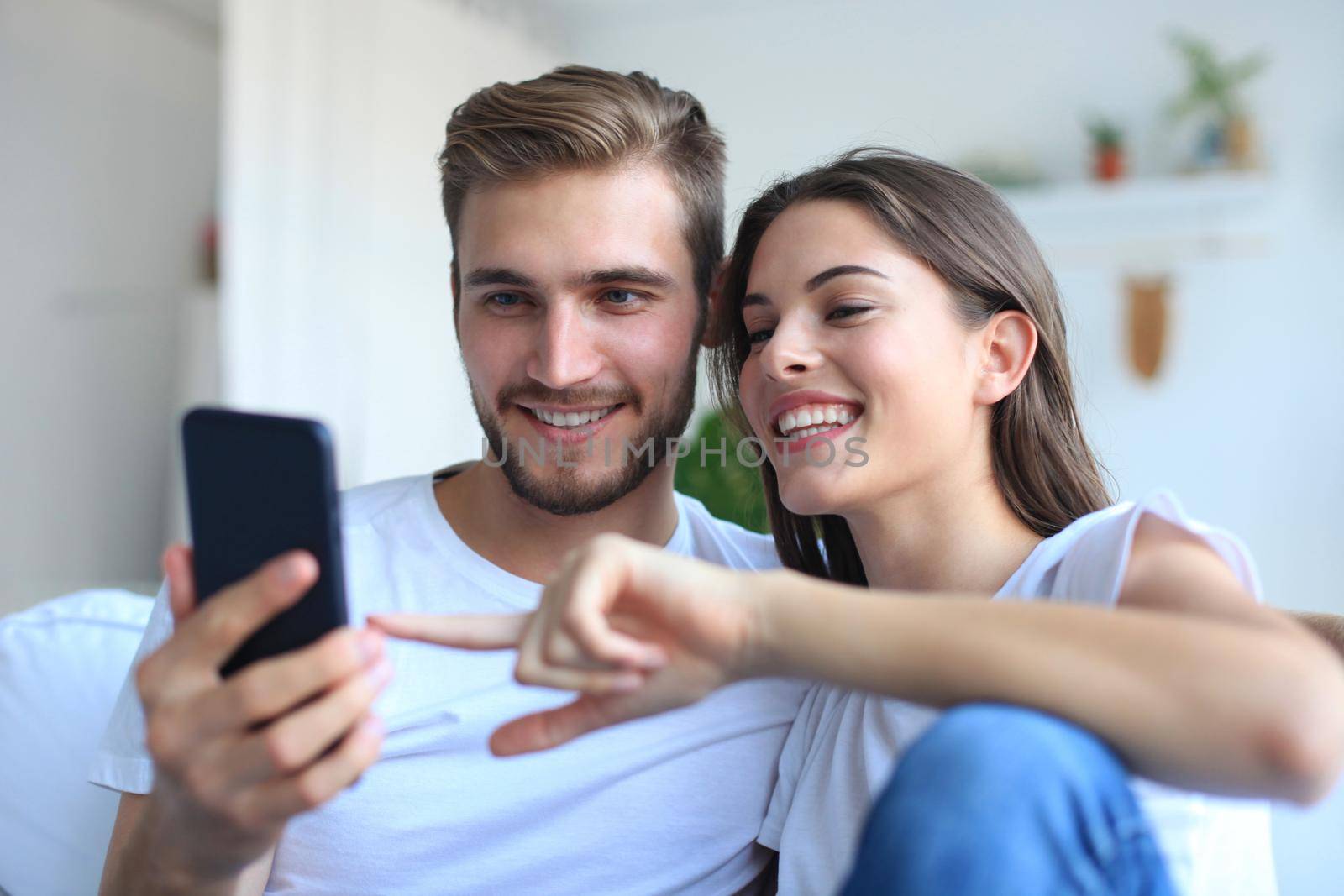 Young couple watching online content in a smart phone sitting on a sofa at home in the living room.