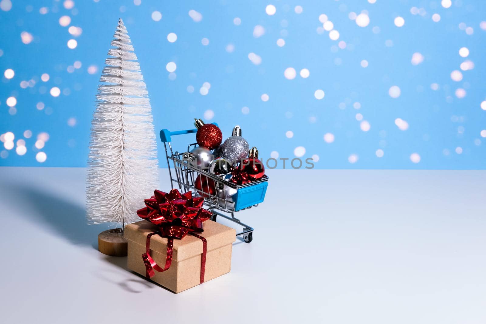 Beautiful Christmas or New Year banner with copy space. Toy Christmas tree, gift box with red bow and shopping trolley full of Christmas baubles on blue background with blurred lights.
