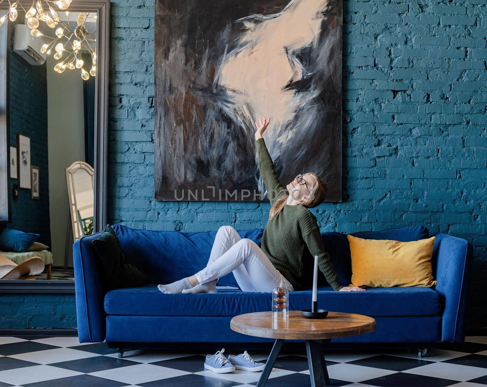 Young caucasian woman in green sweater and white jeans lying on sofa in stylish blue living room, using mobile
