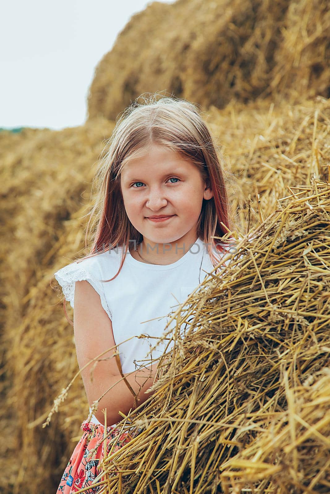 A blonde schoolgirl in a pink dress climbed on large bales of straw on a hot summer day