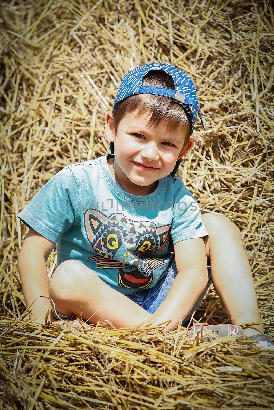A boy in a blue baseball cap sits on a haystack and smiles on a hot summer day