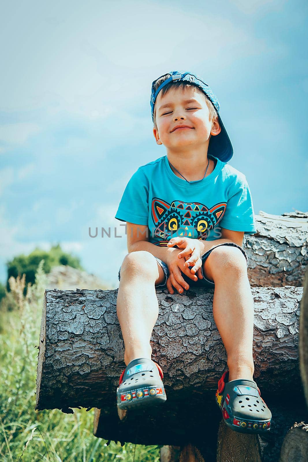 A little boy in a blue baseball cap and denim shorts sits on a log and thinks about something