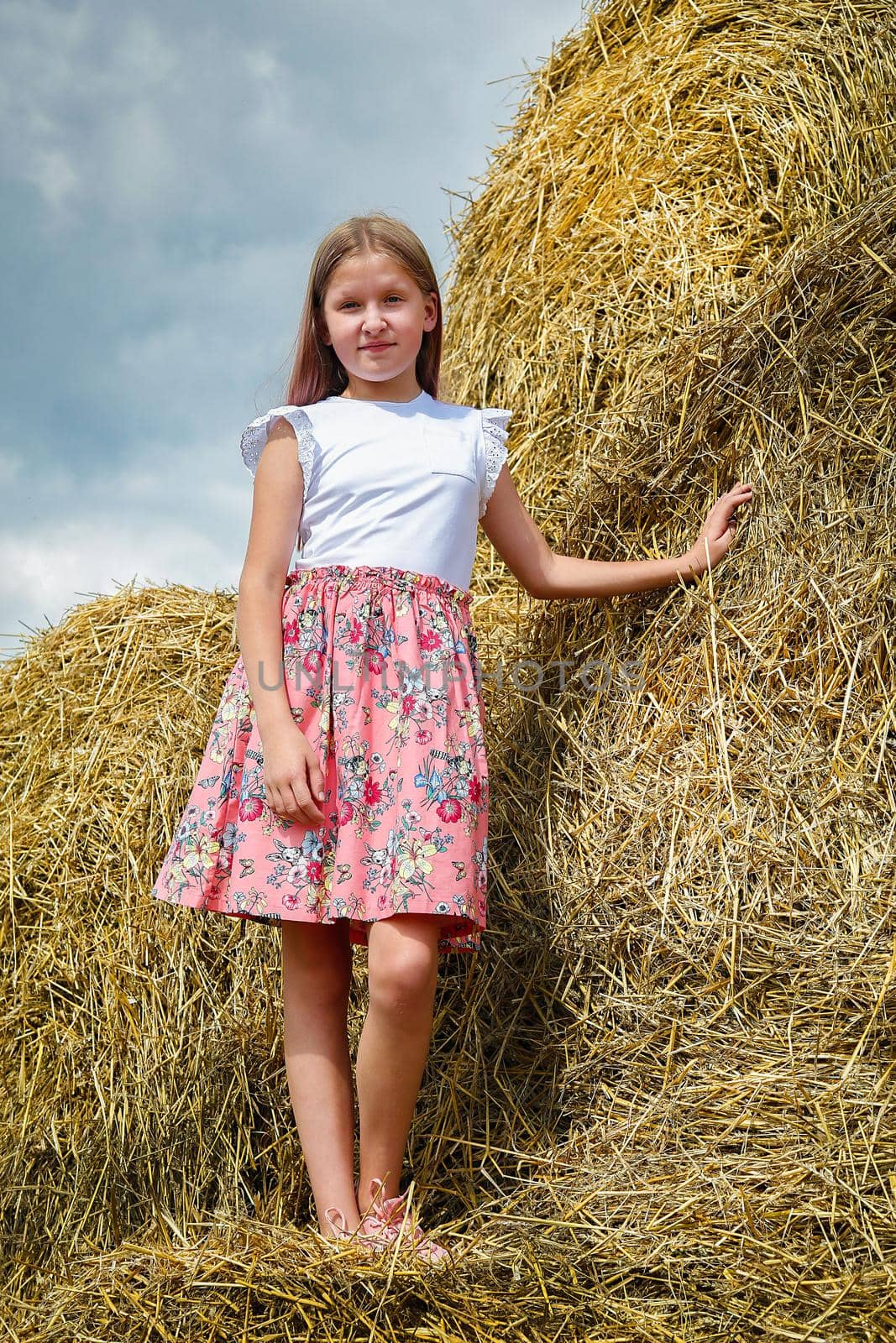 Model Girl with long blonde hair in a pink dress climbed on large bales of straw on a hot summer day