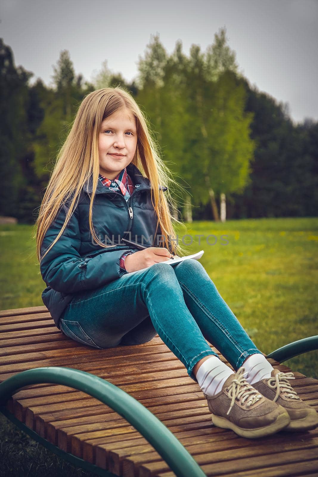 Blonde girl writes a letter sitting on a wooden chaise longue in the Park