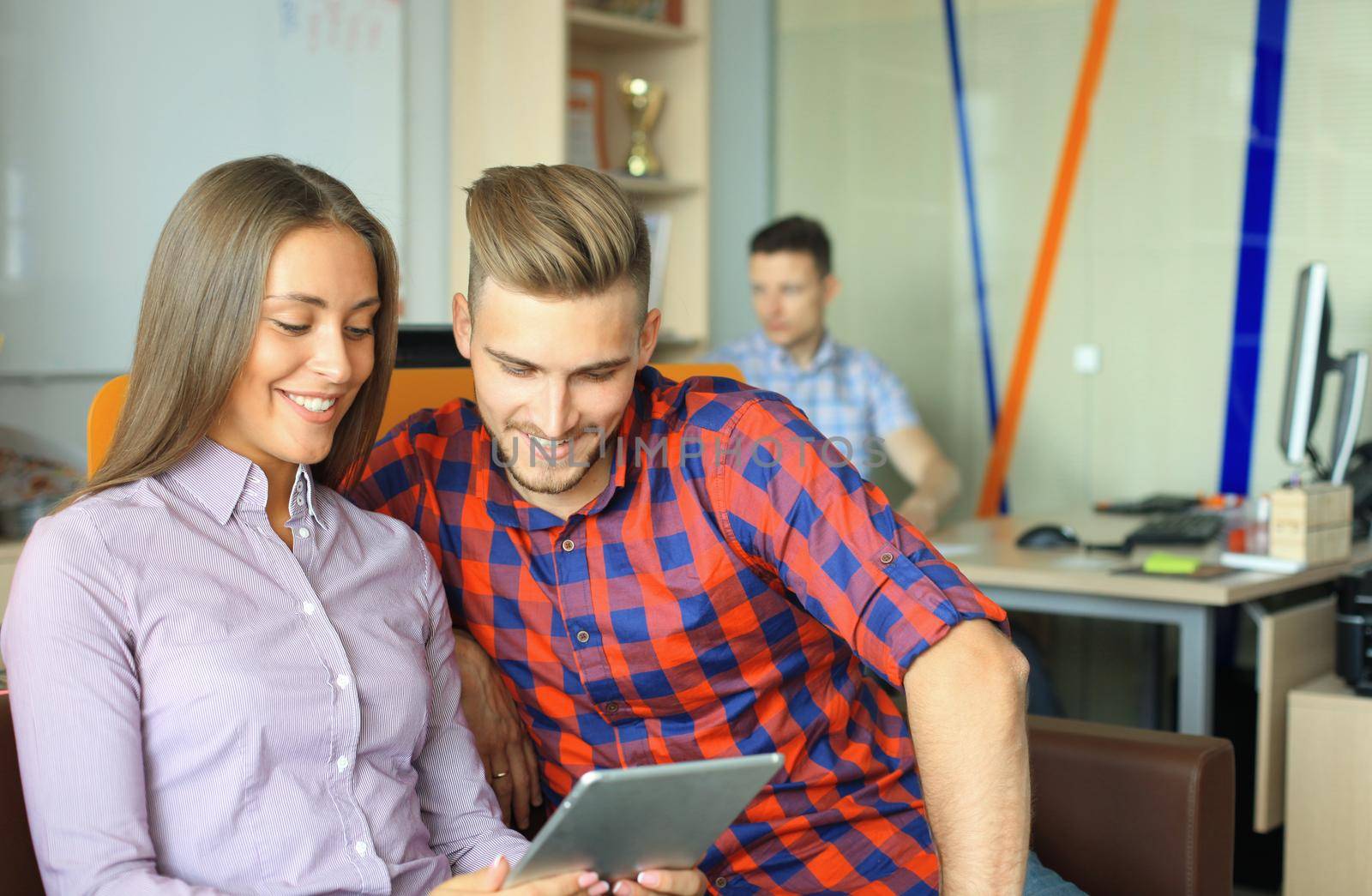 Two confident young people looking at touchpad while their colleagues working in the background