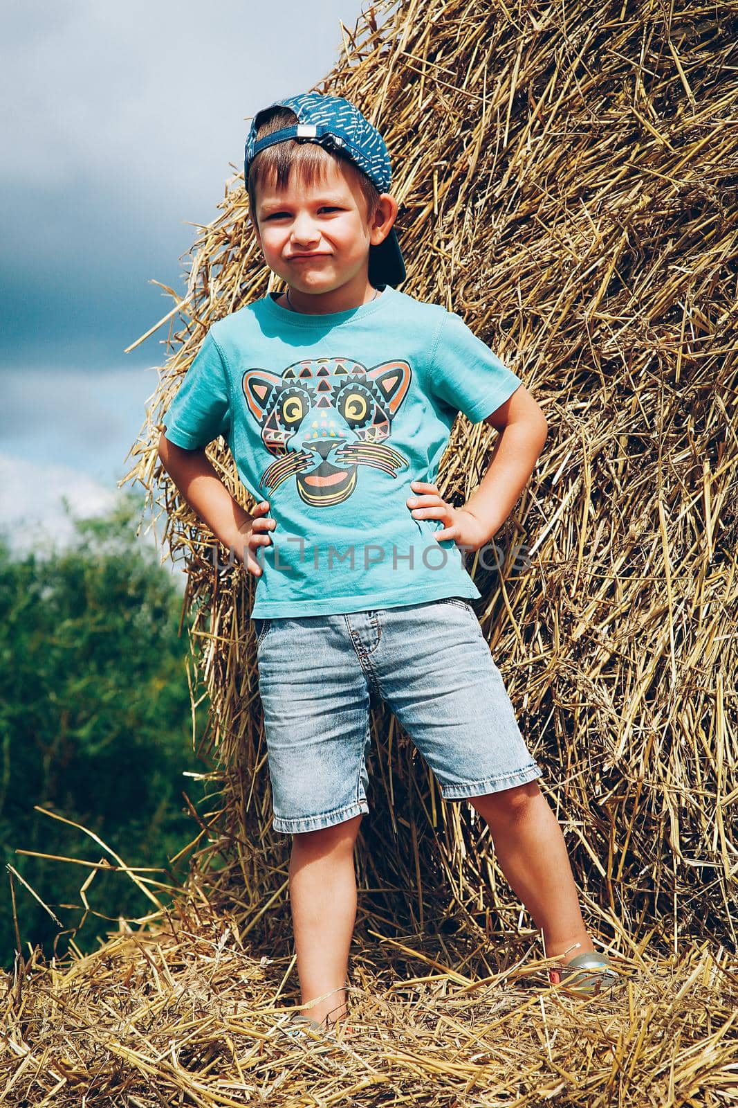 A little boy in a blue baseball cap and short denim shorts plays on straw bales on a hot summer day