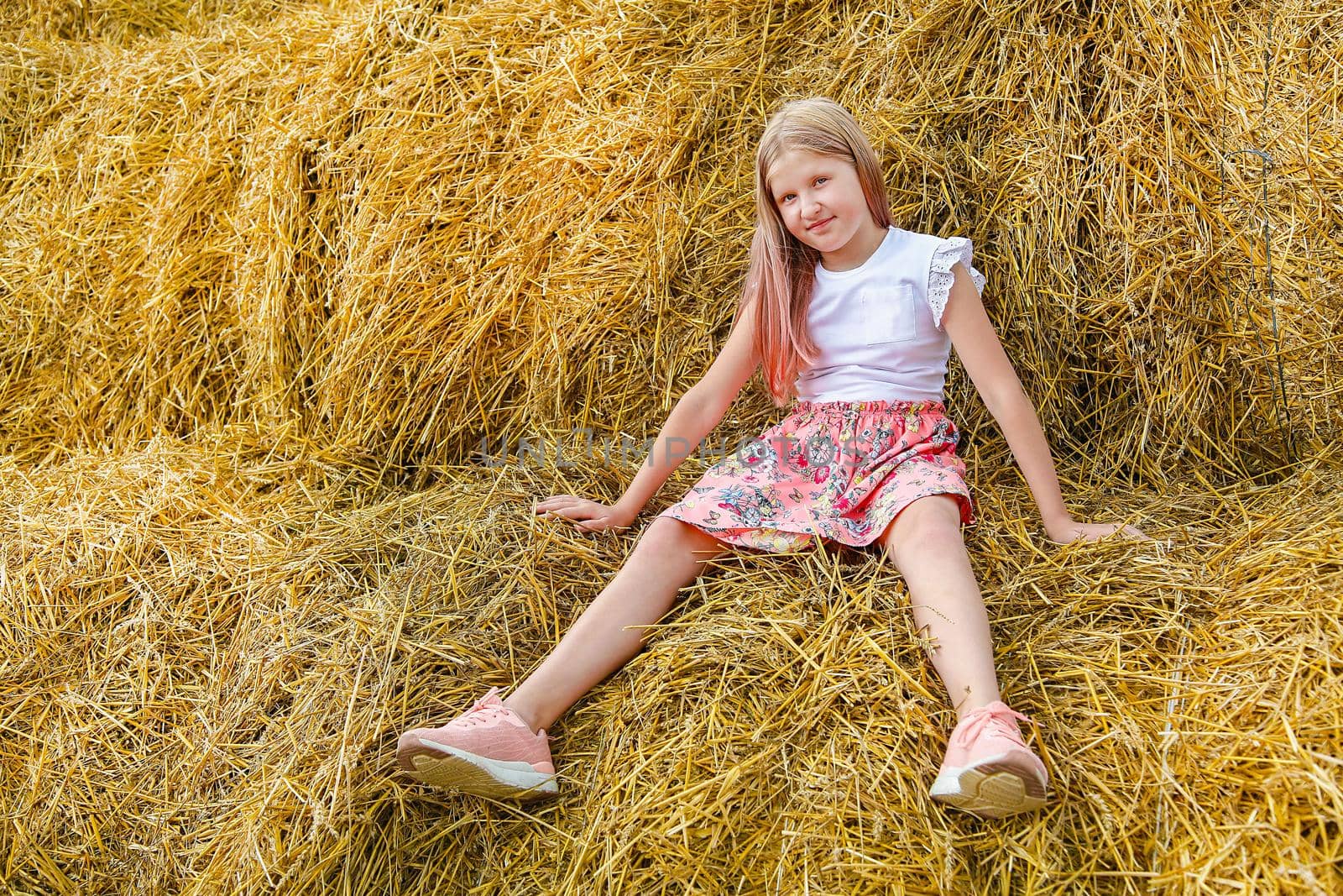 A blonde girl with long hair in a pink dress sits on a bale of straw on a hot summer day like a doll