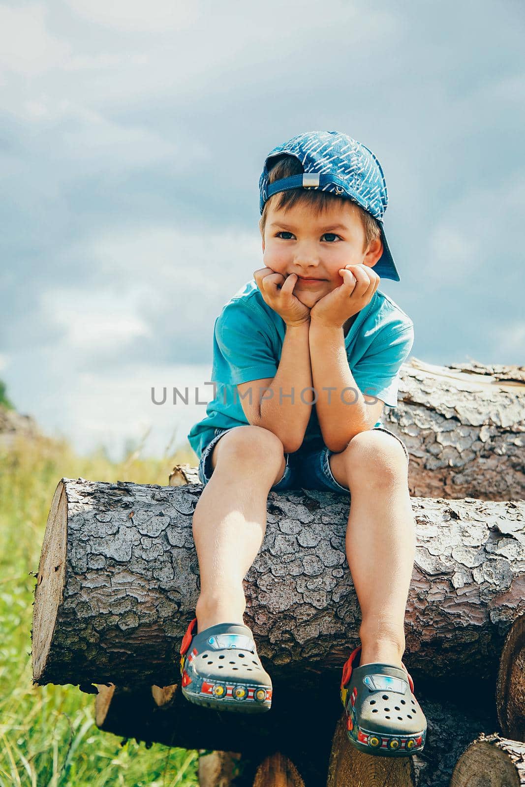 A small child in a blue baseball cap and short denim shorts sits on a log and laughs merrily
