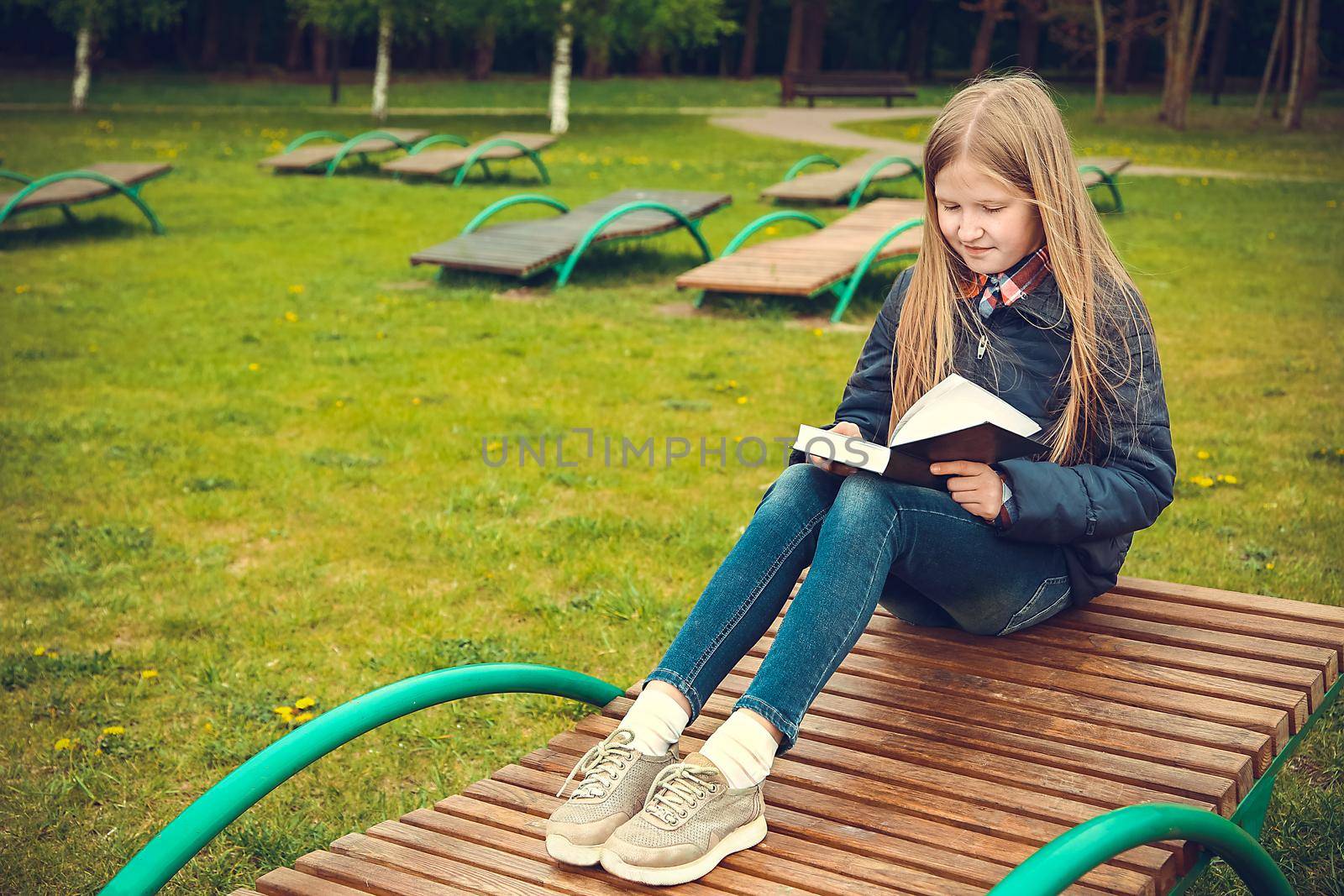 School girl with blond hair sitting on a wooden bench in a forested area and reading a book