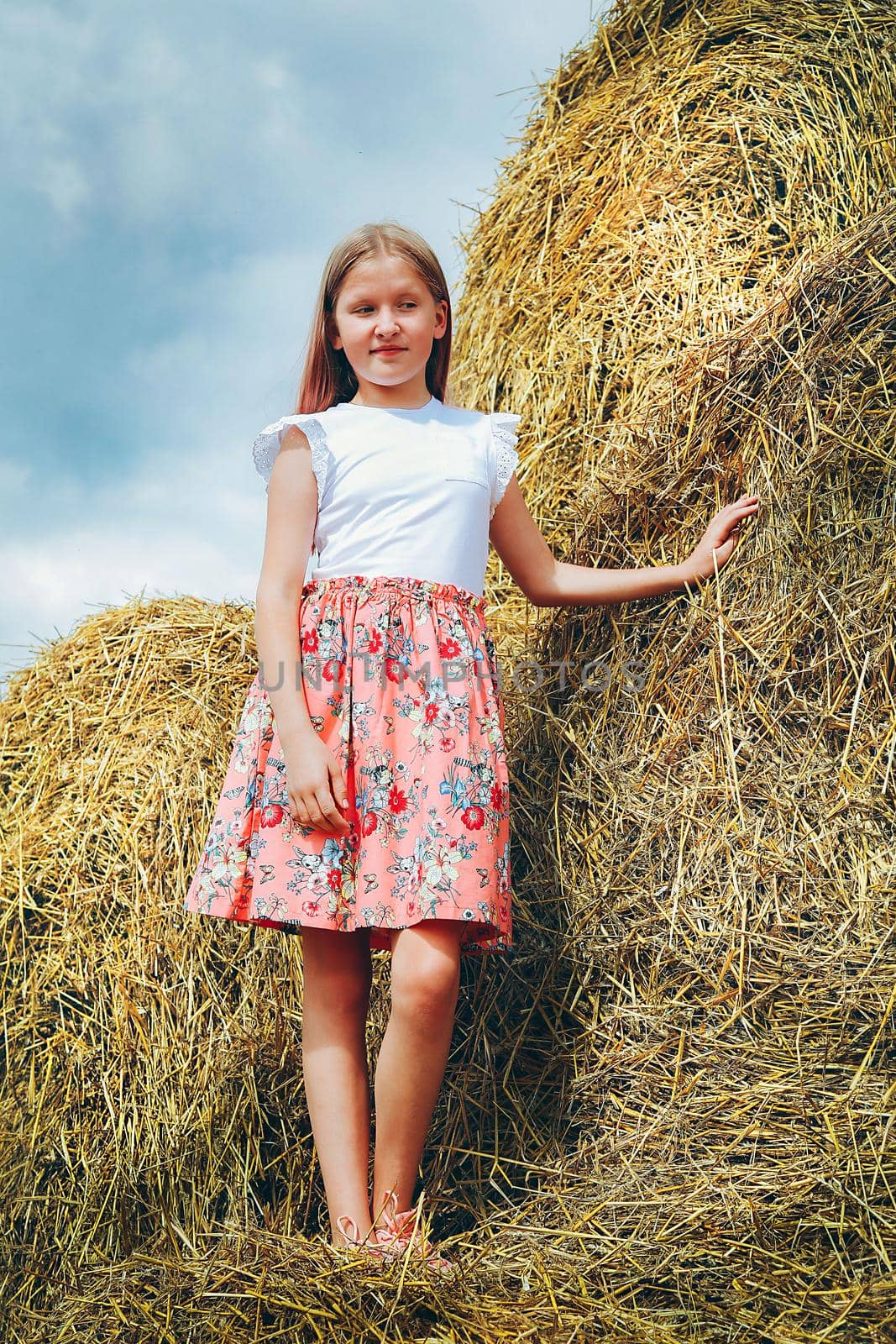 A schoolgirl with long hair in a pink dress climbed on large bales of straw on a hot summer day