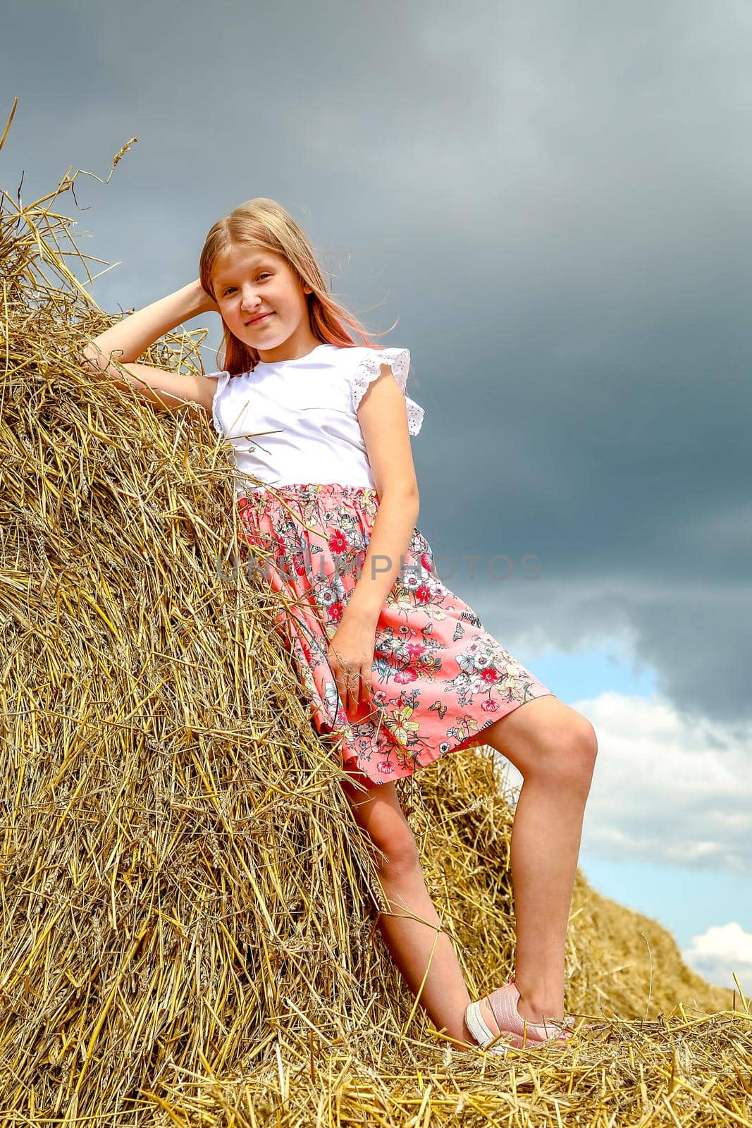 A beautiful blonde girl in a pink dress climbed on large bales of straw on a summer day