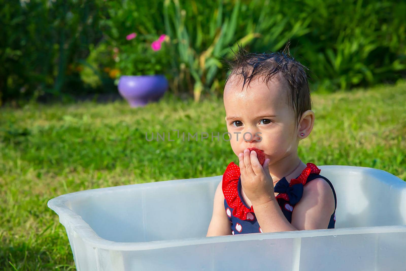 A little girl in a swimsuit sitting in a white bath and eating strawberries