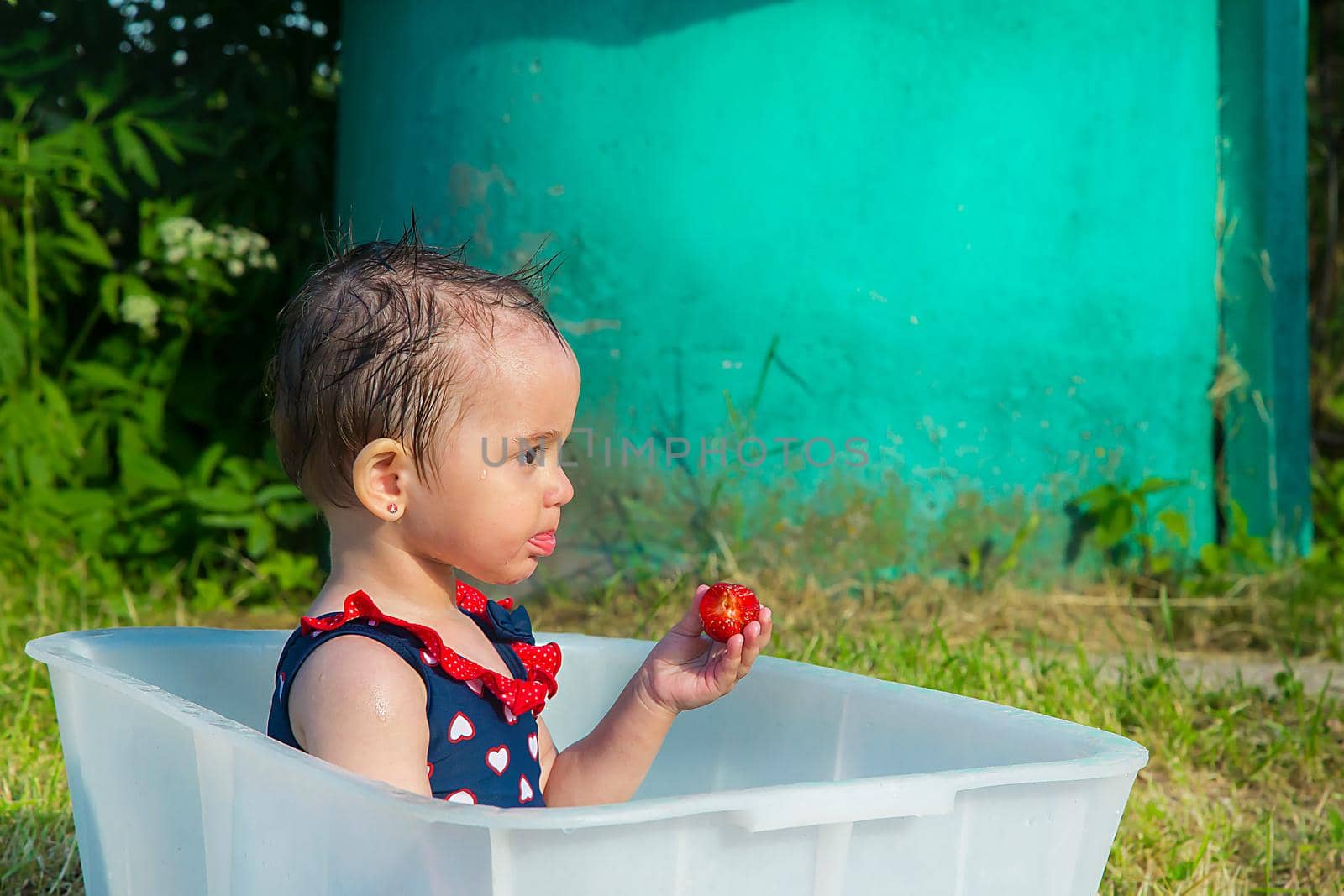 A little girl in a swimsuit sitting in a bath eating ripe strawberries by Mastak80