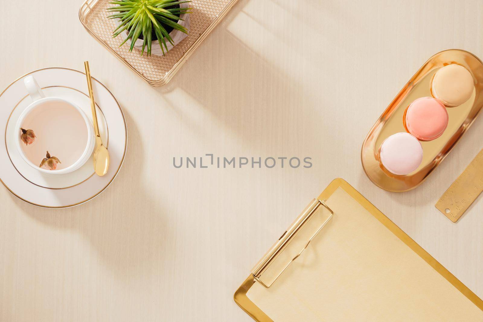 Modern gold stylized home office desk with folder, macaroons, coffee mug on beige background. Flat lay, top view lifestyle concept.`