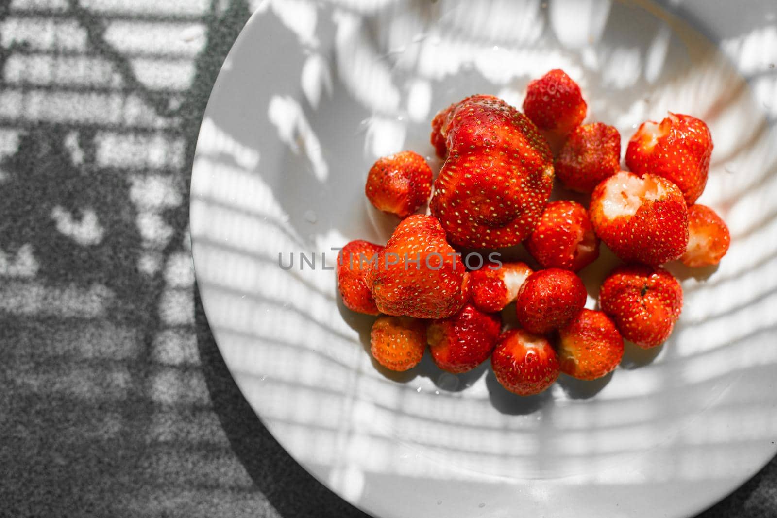 Many juicy fresh ripe red strawberry berries lie in a white ceramic plate on the table under bright sunlight by StudioLucky