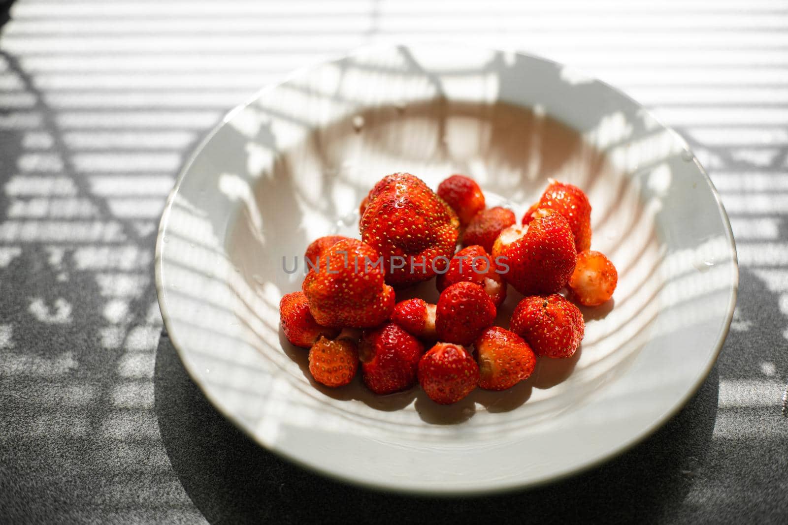 Top view of a ceramic plate full of fresh and sweet organic strawberries picked in the garden in sunlight. Window shade on the kitchen table.
